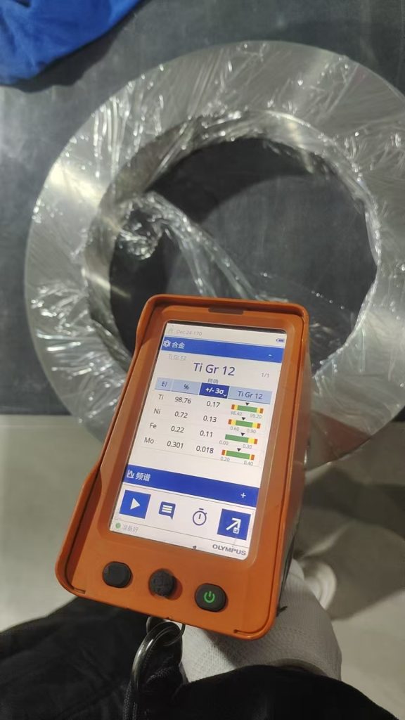 Olympus handheld XRF analyzer performing PMI on a titanium Grade 12 ring, showing the alloy composition readout