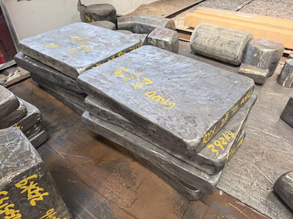 Forged titanium slabs stacked at the works, hand marked in yellow paint with piece and weight identification